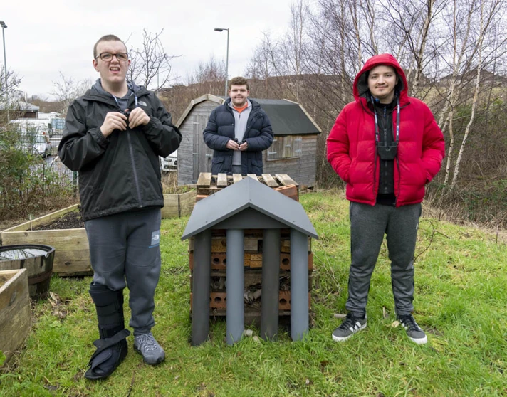 Three young men standing outside beside a handmade bug hotel, surrounded by greenery and raised garden beds. Three young men standing outside beside a handmade bug hotel, surrounded by greenery and raised garden beds.